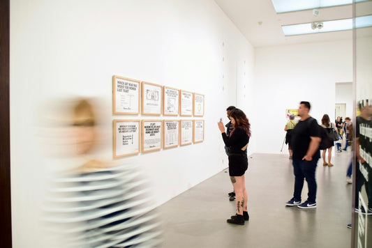 Onlookers in an art gallery. There are white walls with images framed on the walls. A person in the foreground is blurred indicating movement. 